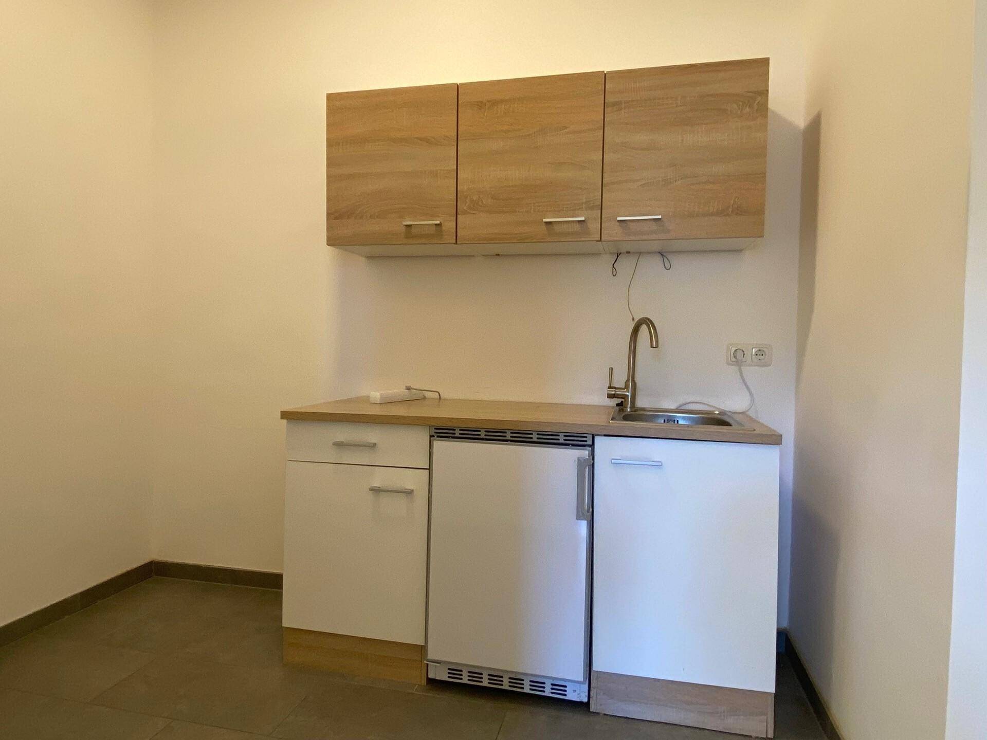 Kompakte Küchenzeile mit Holzschränken, Spüle und integriertem Kühlschrank auf Fliesenboden.
