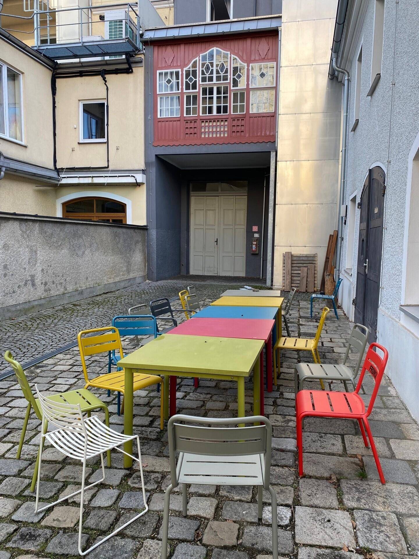 Gemütlicher Innenhof mit bunten Tischen und Stühlen auf Kopfsteinpflaster, ideal für entspannte Pausen im Freien.