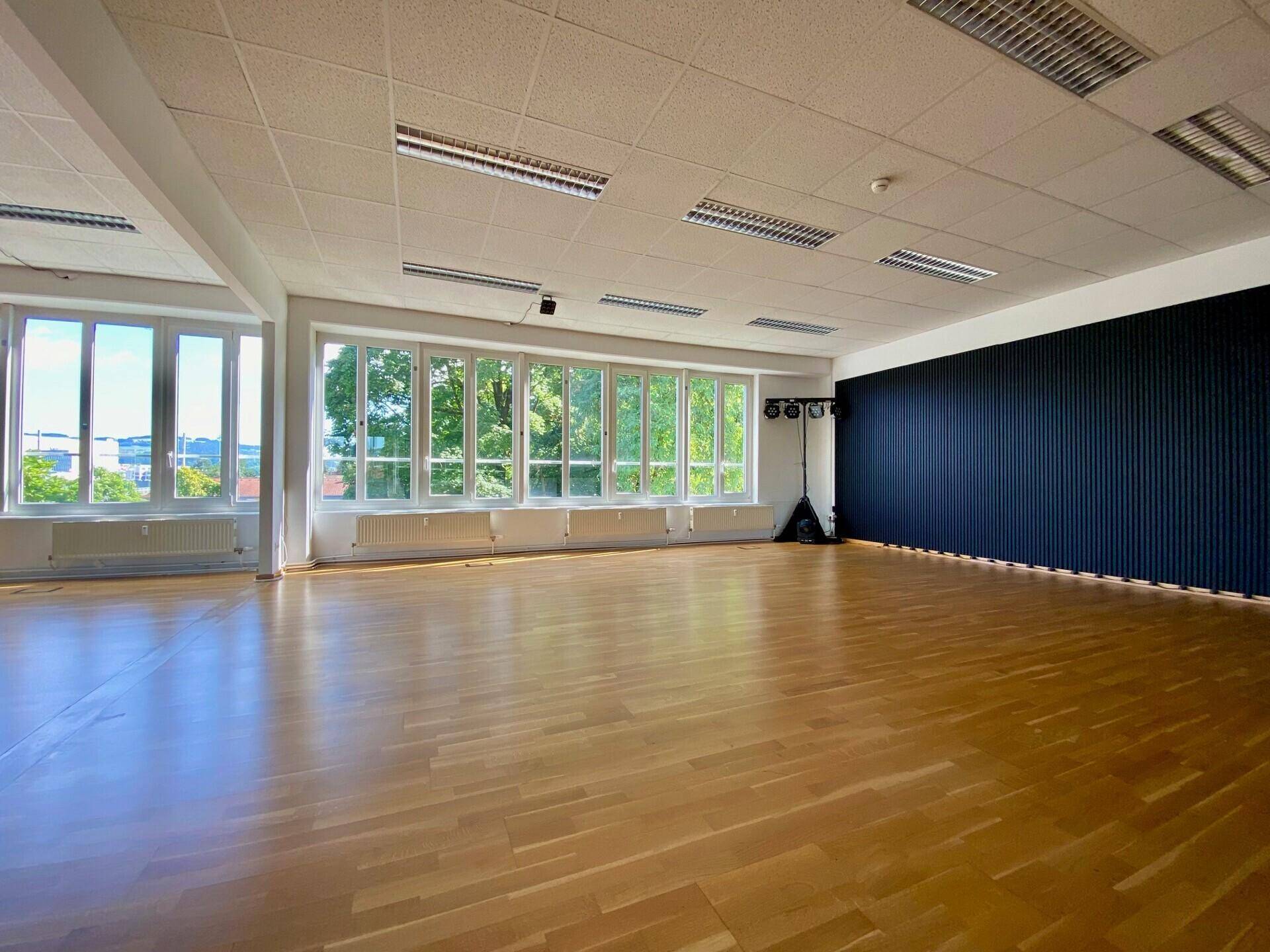 Heller Studiobereich mit Holzboden, großen Fenstern und einer markanten schwarzen Wandverkleidung.