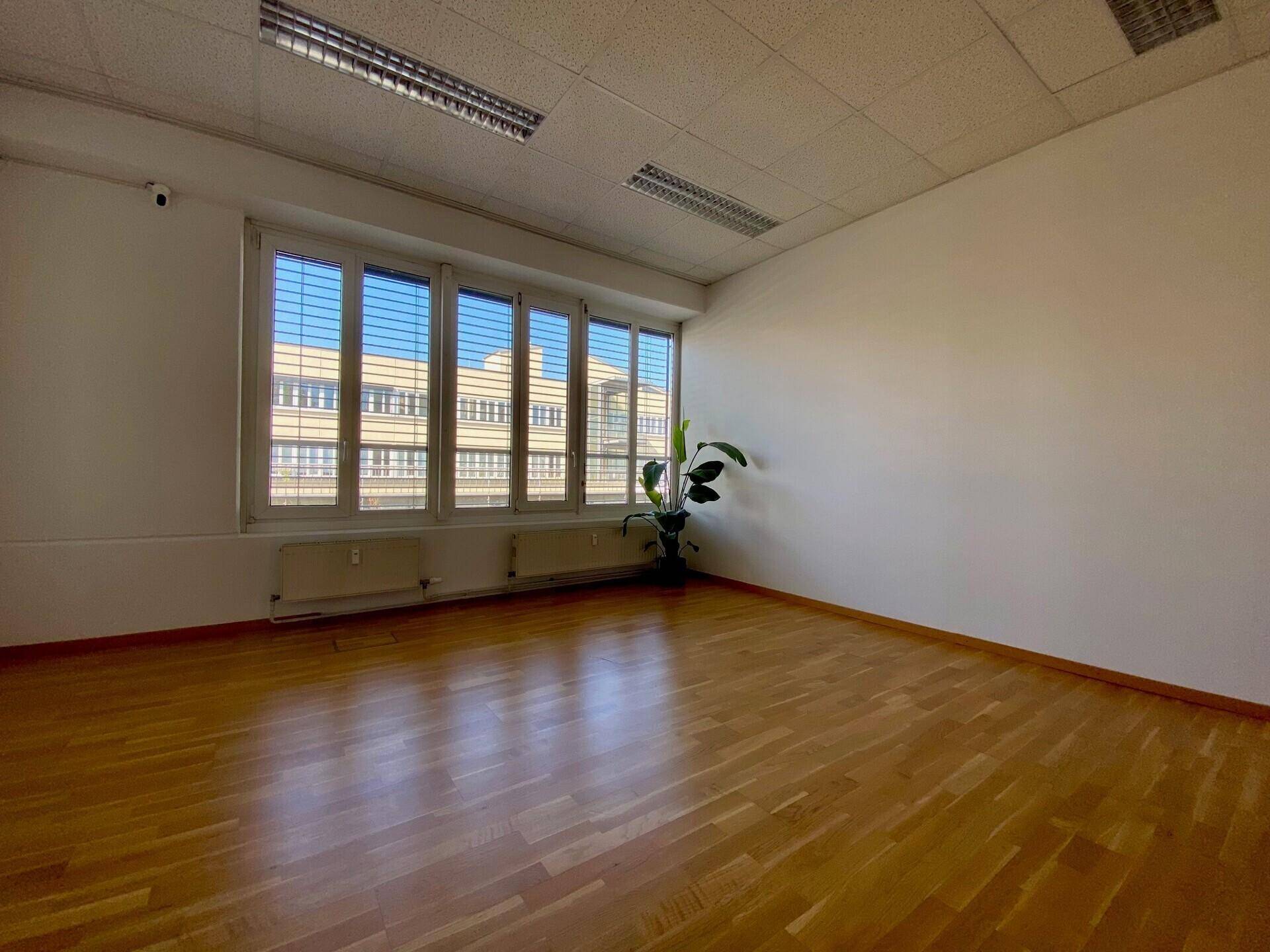 Leerer Raum mit Holzboden und einer Fensterfront mit Jalousien, die einen Blick auf ein Gebäude bietet.