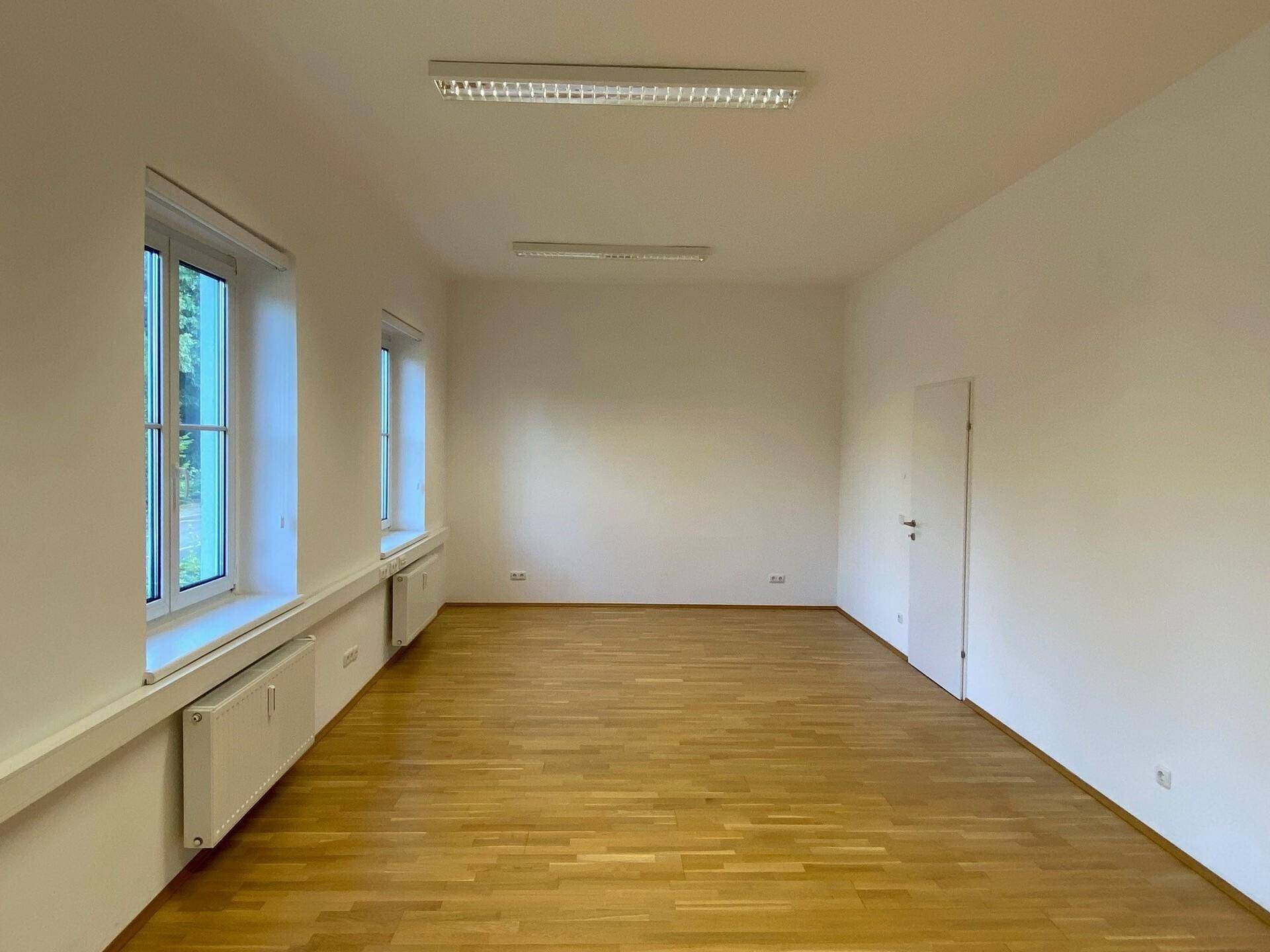 Langer, heller Büroraum mit Parkettboden, weißen Wänden und drei großen Fenstern.
