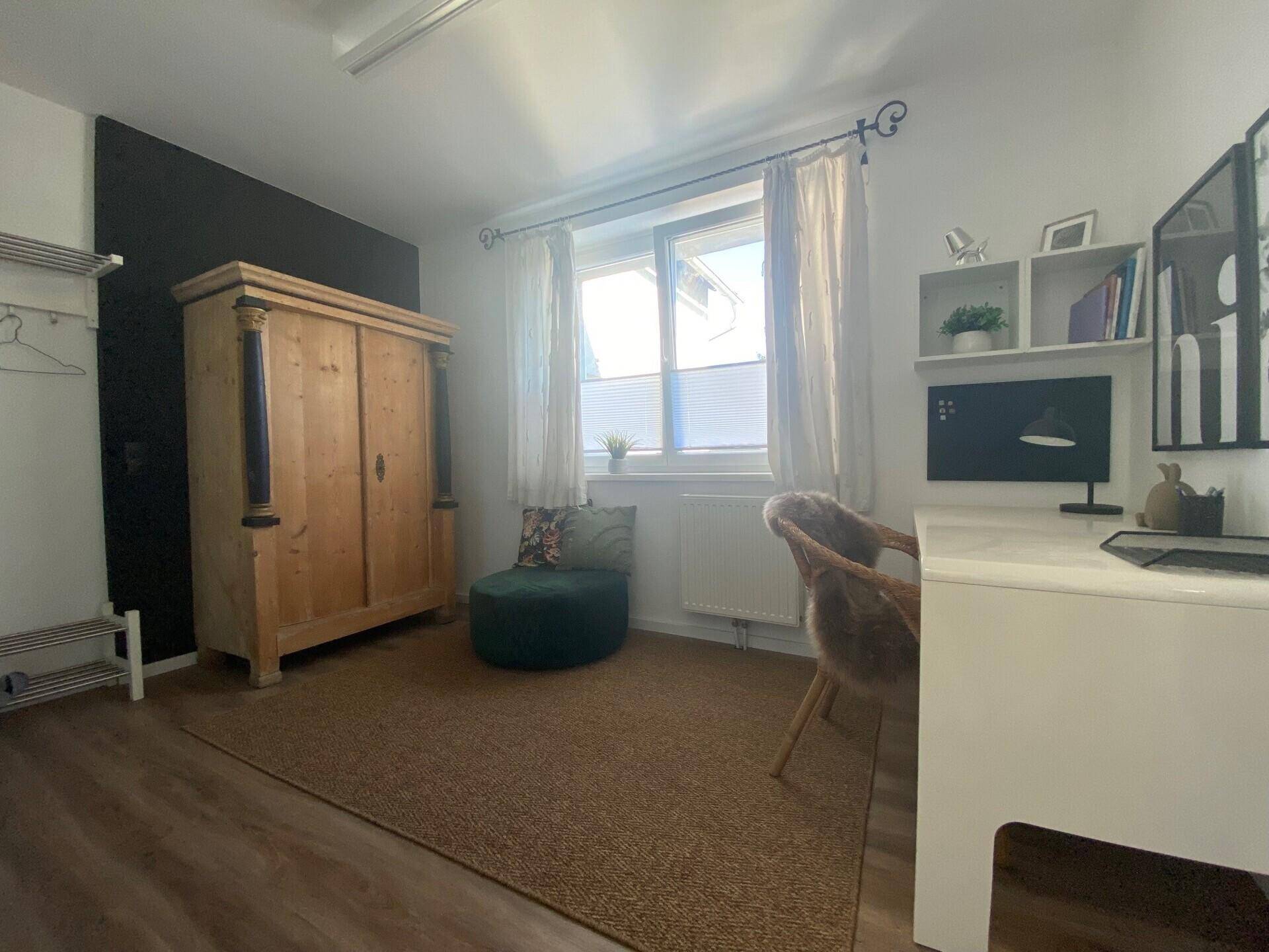 Arbeitszimmer mit Holzboden, großem Holzschrank und einem Fenster, das viel Tageslicht hereinlässt.