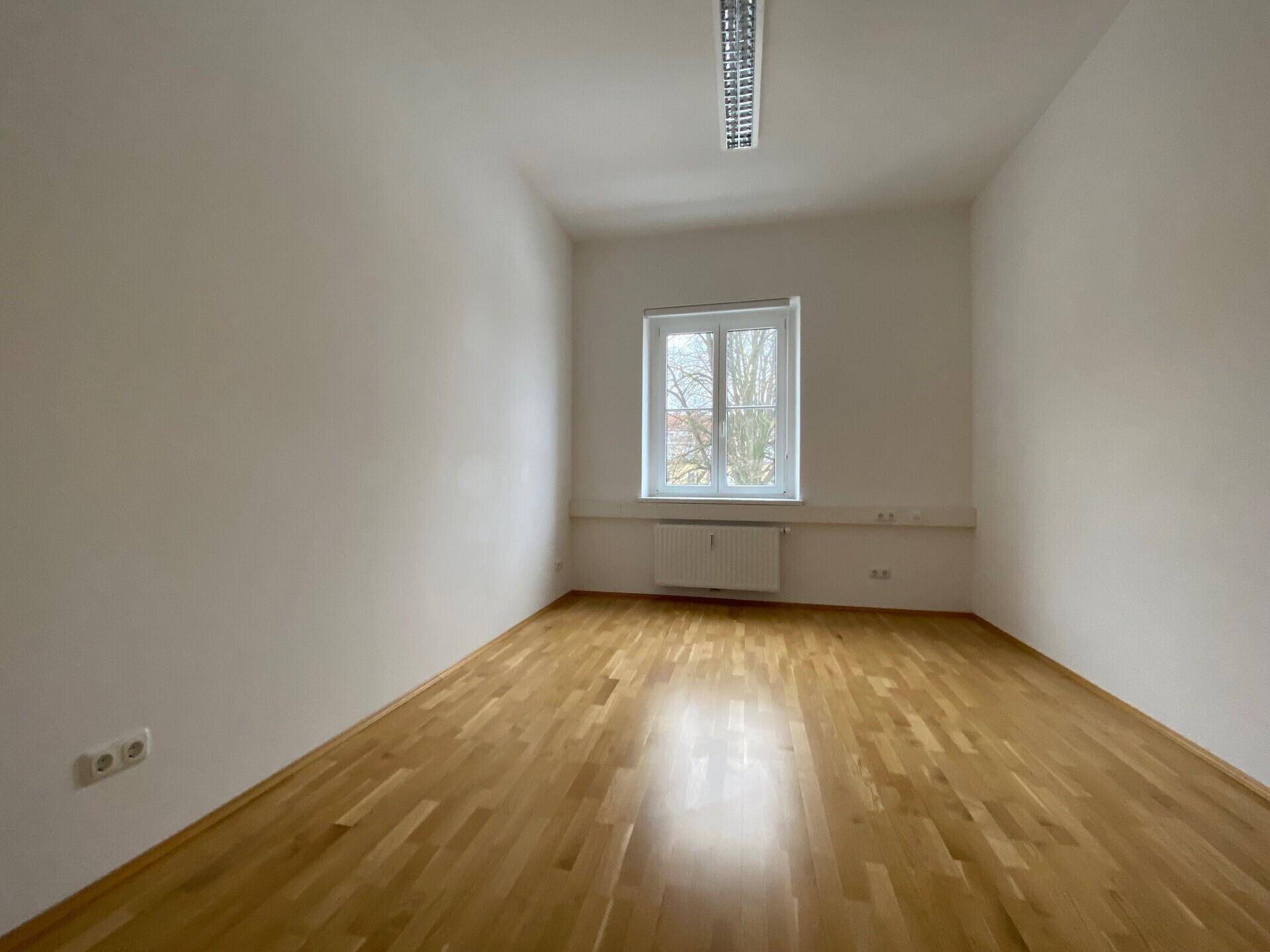 Kleinerer Büroraum mit einem Fenster, das den Blick nach draußen freigibt, und hellem Parkettboden.