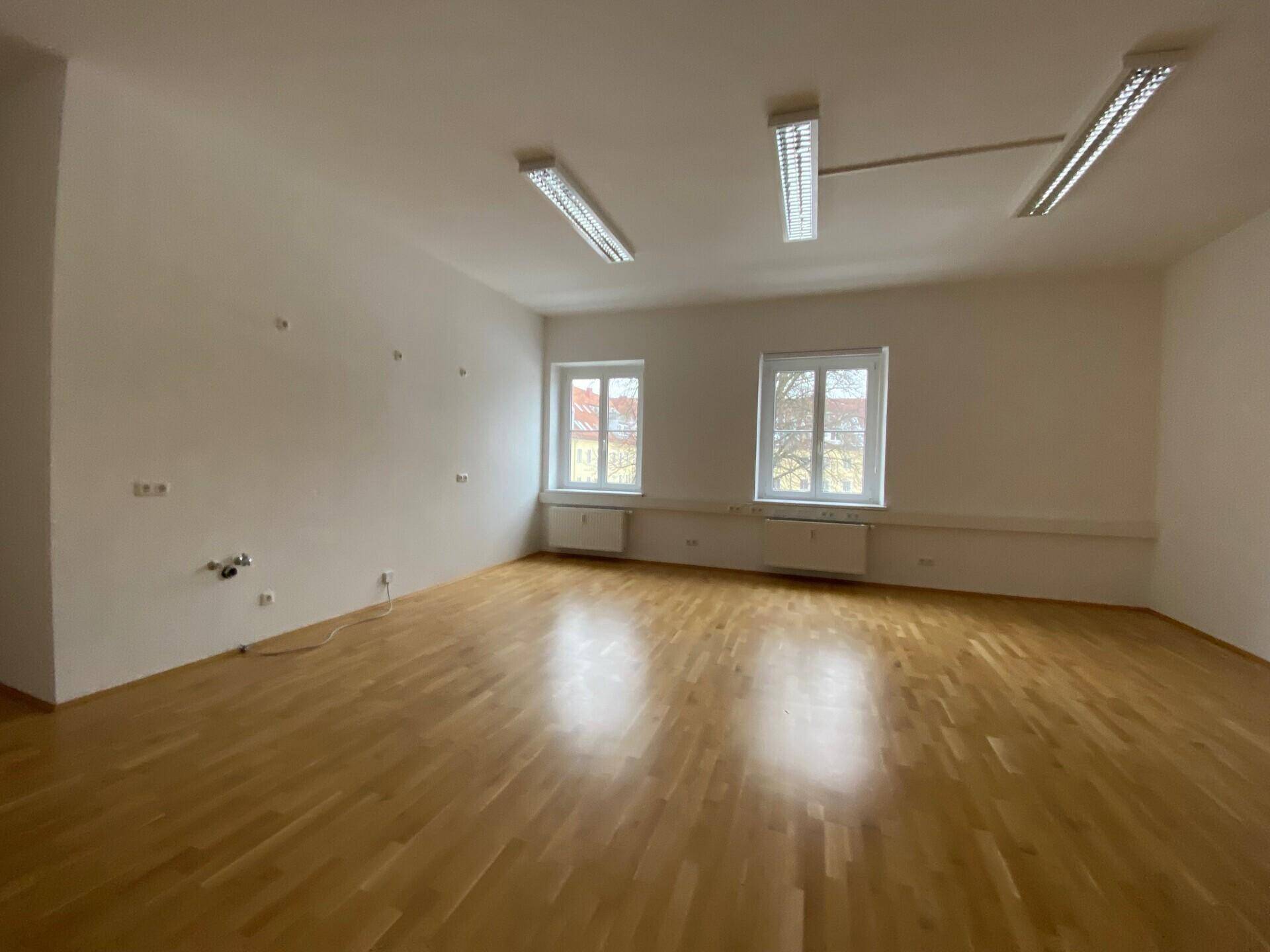 Heller Büroraum mit Holzboden und zwei Fenstern, die eine angenehme Arbeitsatmosphäre schaffen.