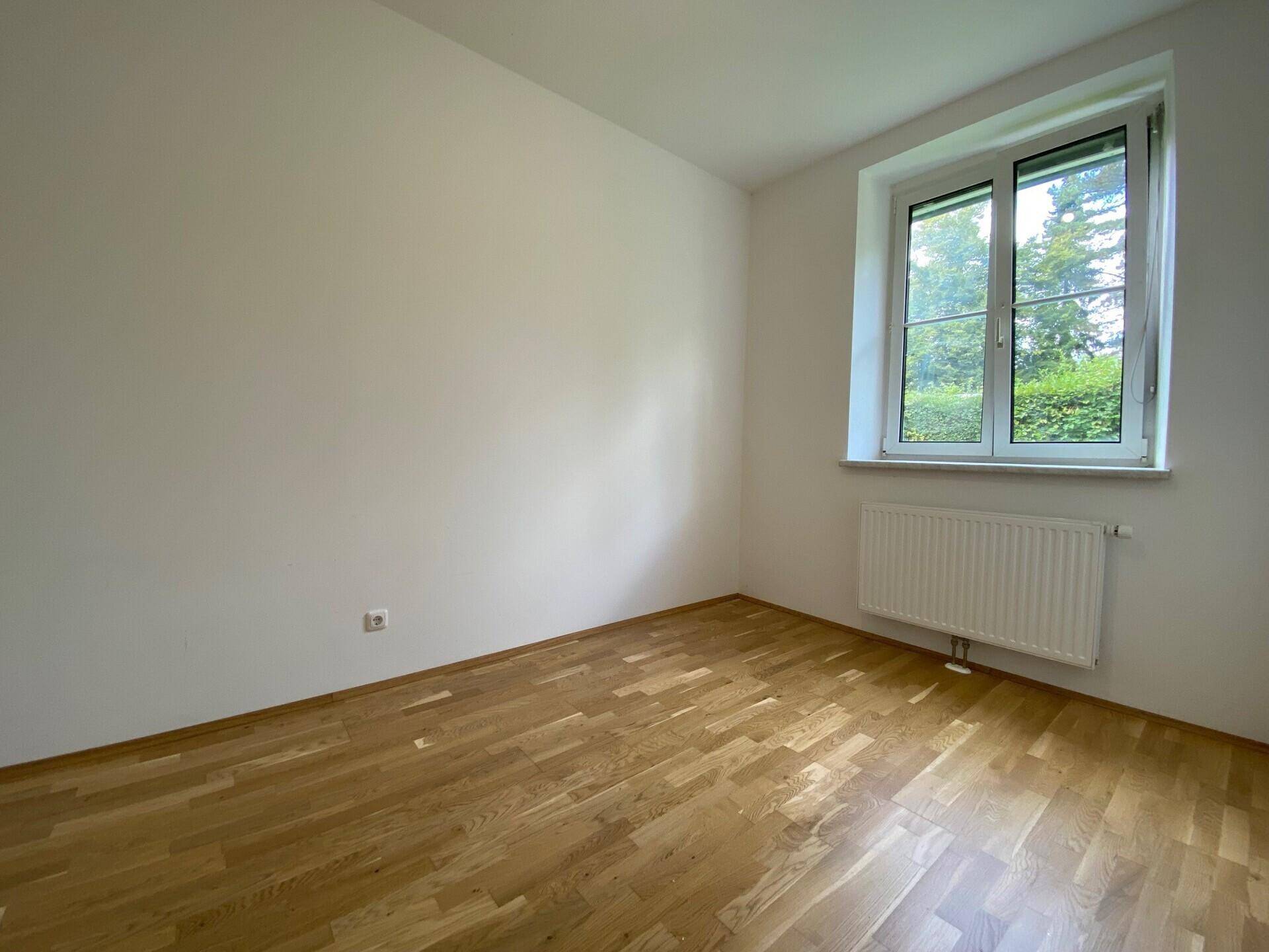 Helles Zimmer mit Parkettboden und großem Fenster, das viel Tageslicht hereinlässt und eine grüne Aussicht bietet.