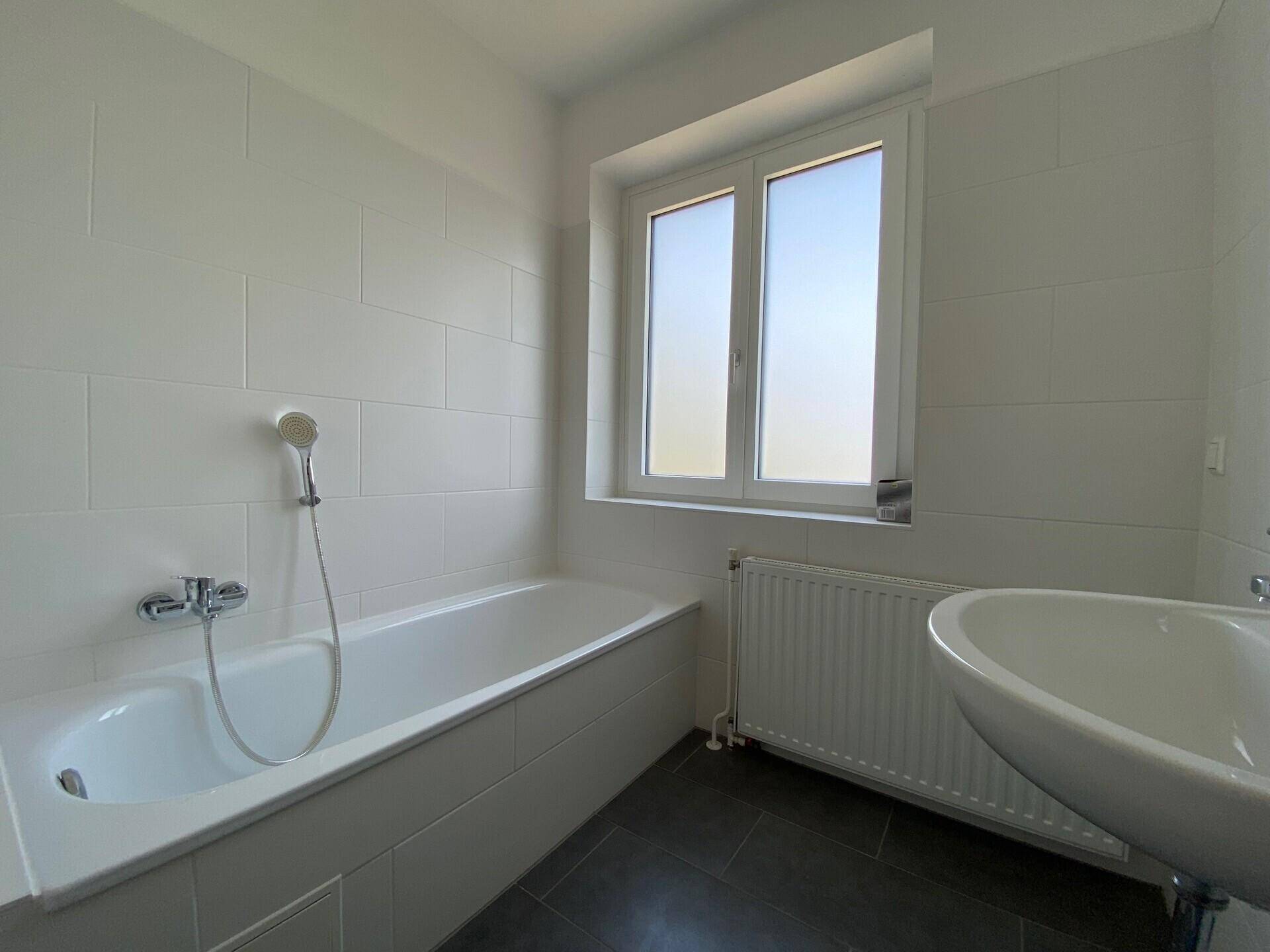 Helles Badezimmer mit Badewanne, Waschbecken, Fenster und modernen Fliesen.