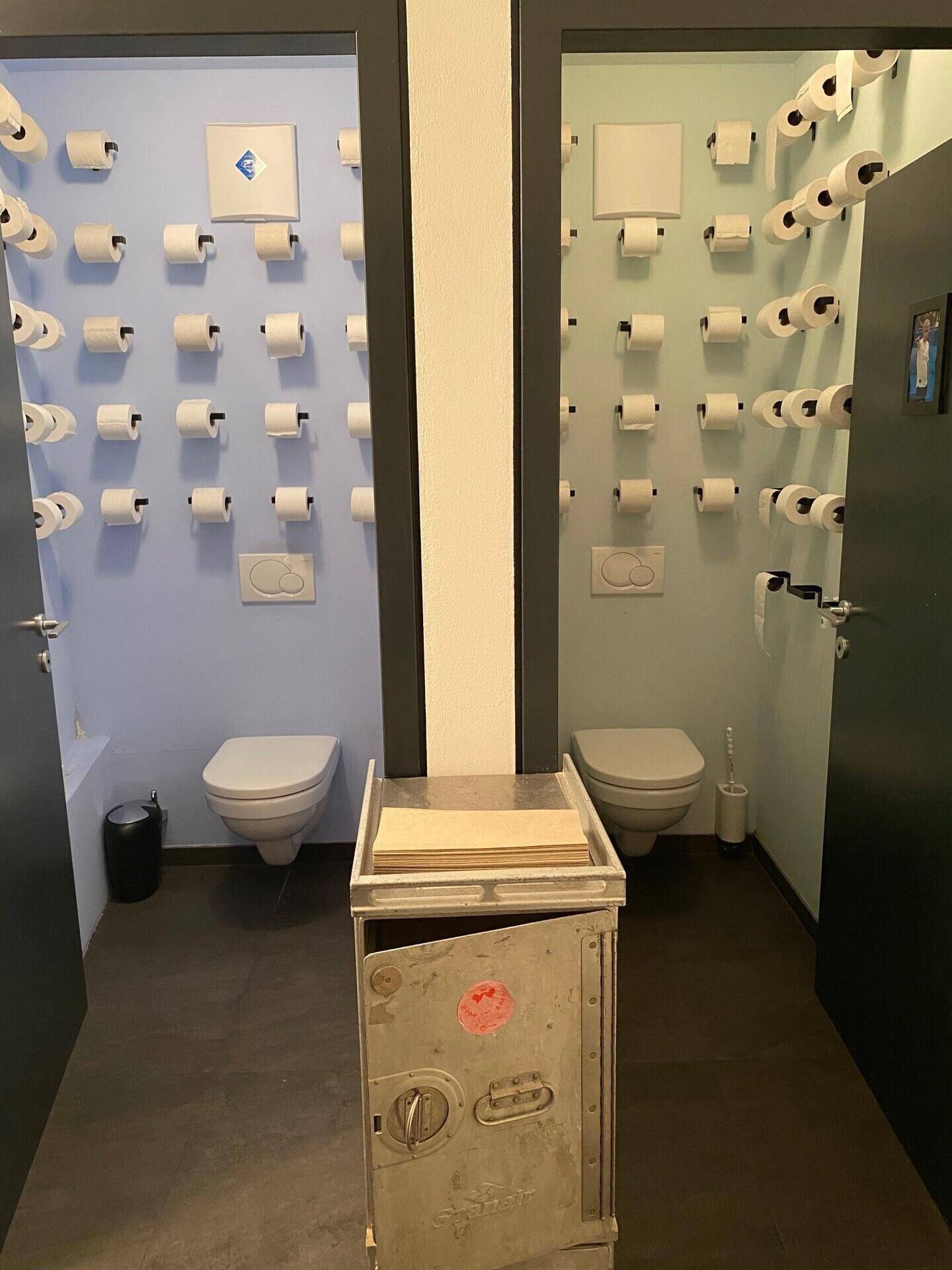 Zwei separate Toilettenräume mit kreativer Wandgestaltung aus vielen Toilettenpapierrollen und modernen Wand-WCs.