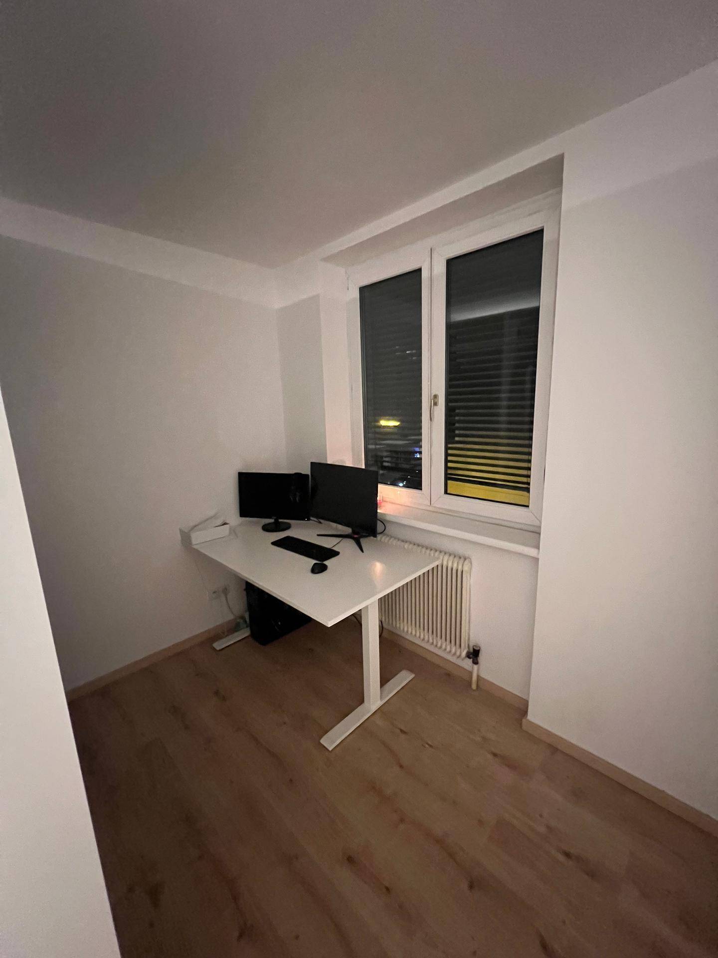 Kleines Arbeitszimmer mit weißem Schreibtisch, zwei Monitoren und Fenster.