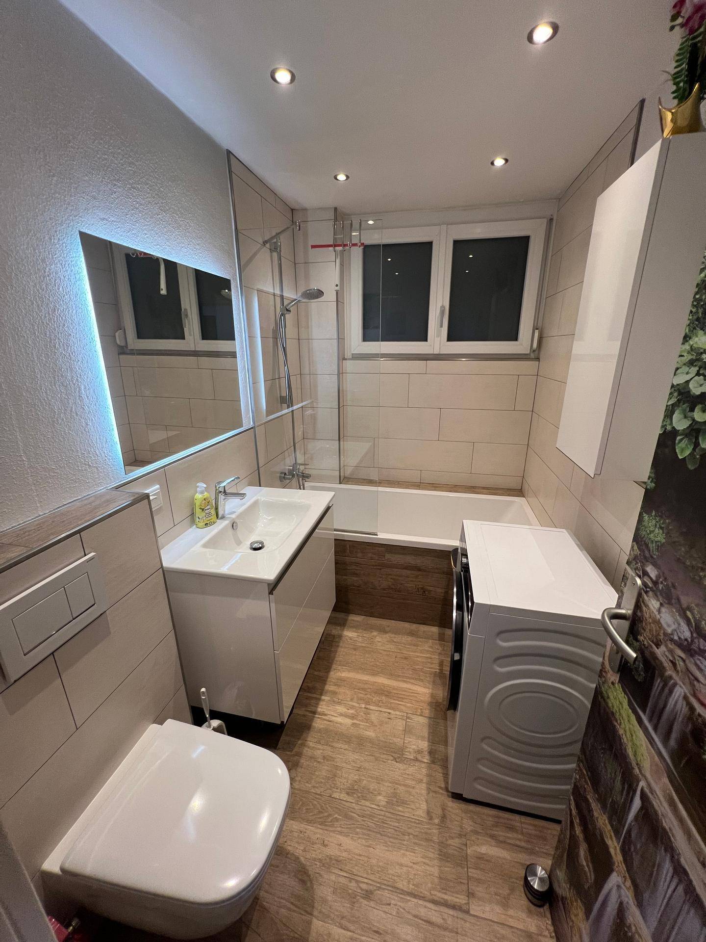 Helles Badezimmer mit Badewanne, Duschabtrennung, Waschbecken und Waschmaschine.