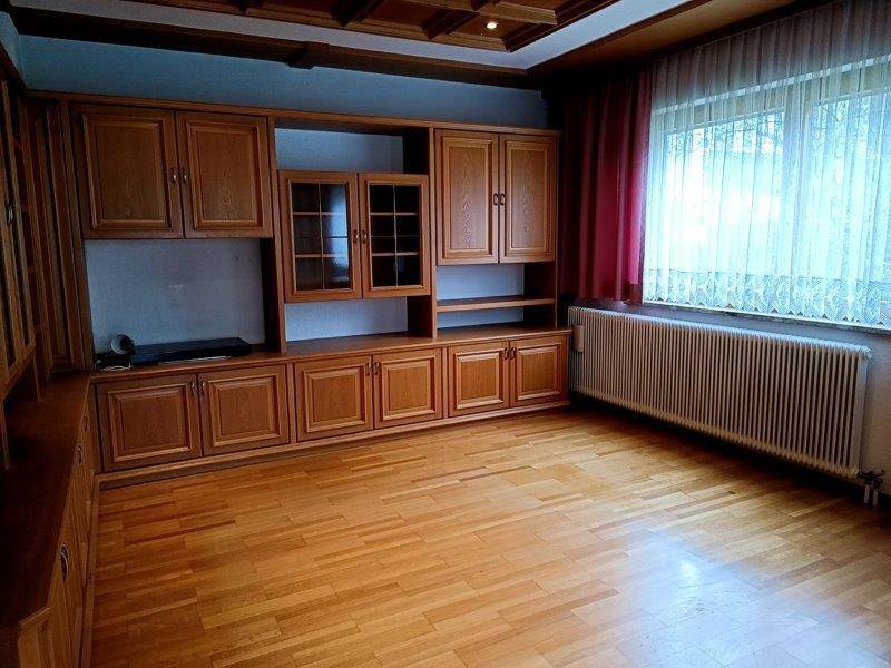 Geräumiges Wohnzimmer mit Holzparkettboden, großer Schrankwand und Fenster mit Vorhängen.