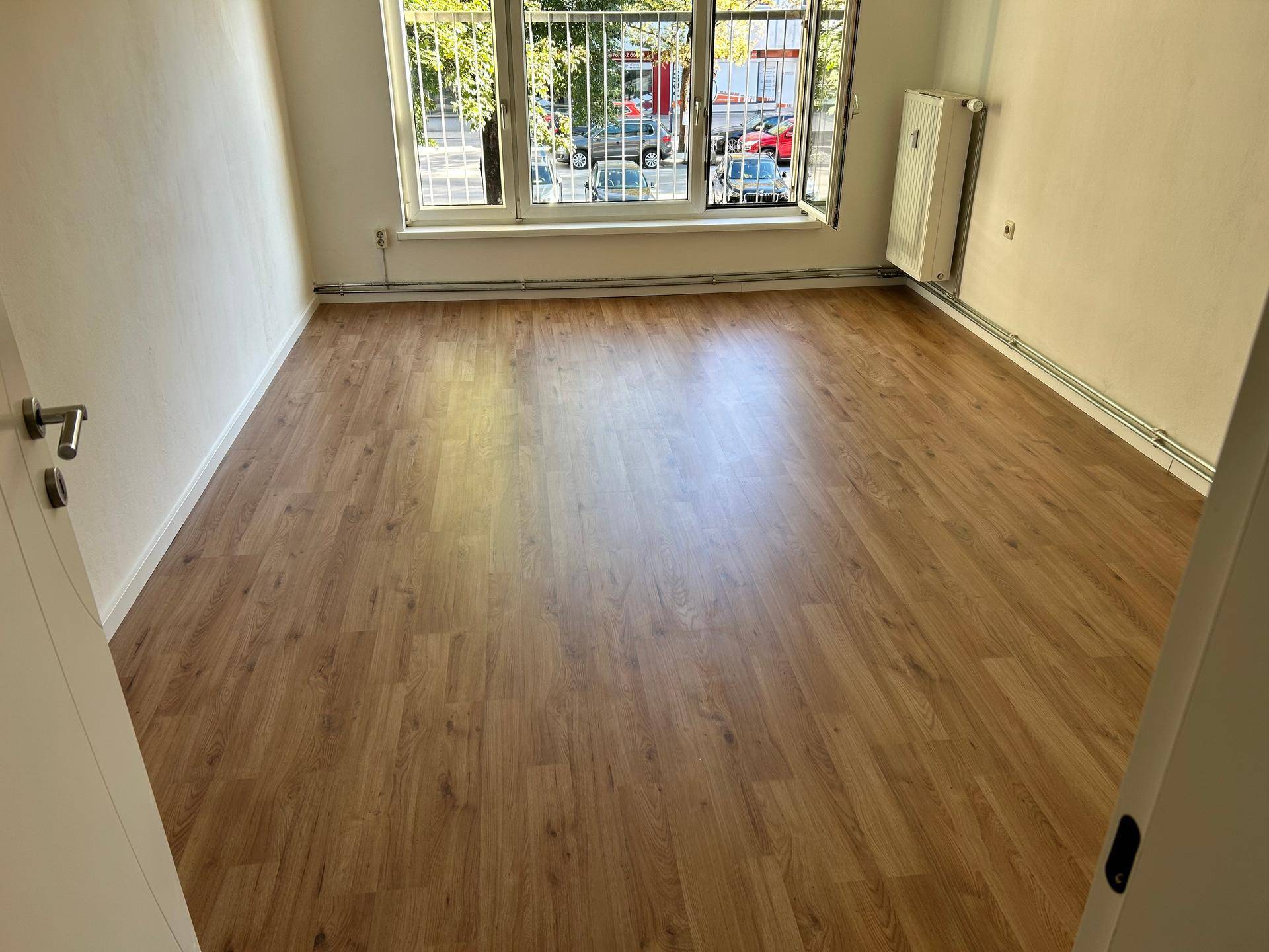 Geräumiges Zimmer mit hellem Laminatboden, Heizkörper und Fenster mit Blick auf Bäume und Straße.