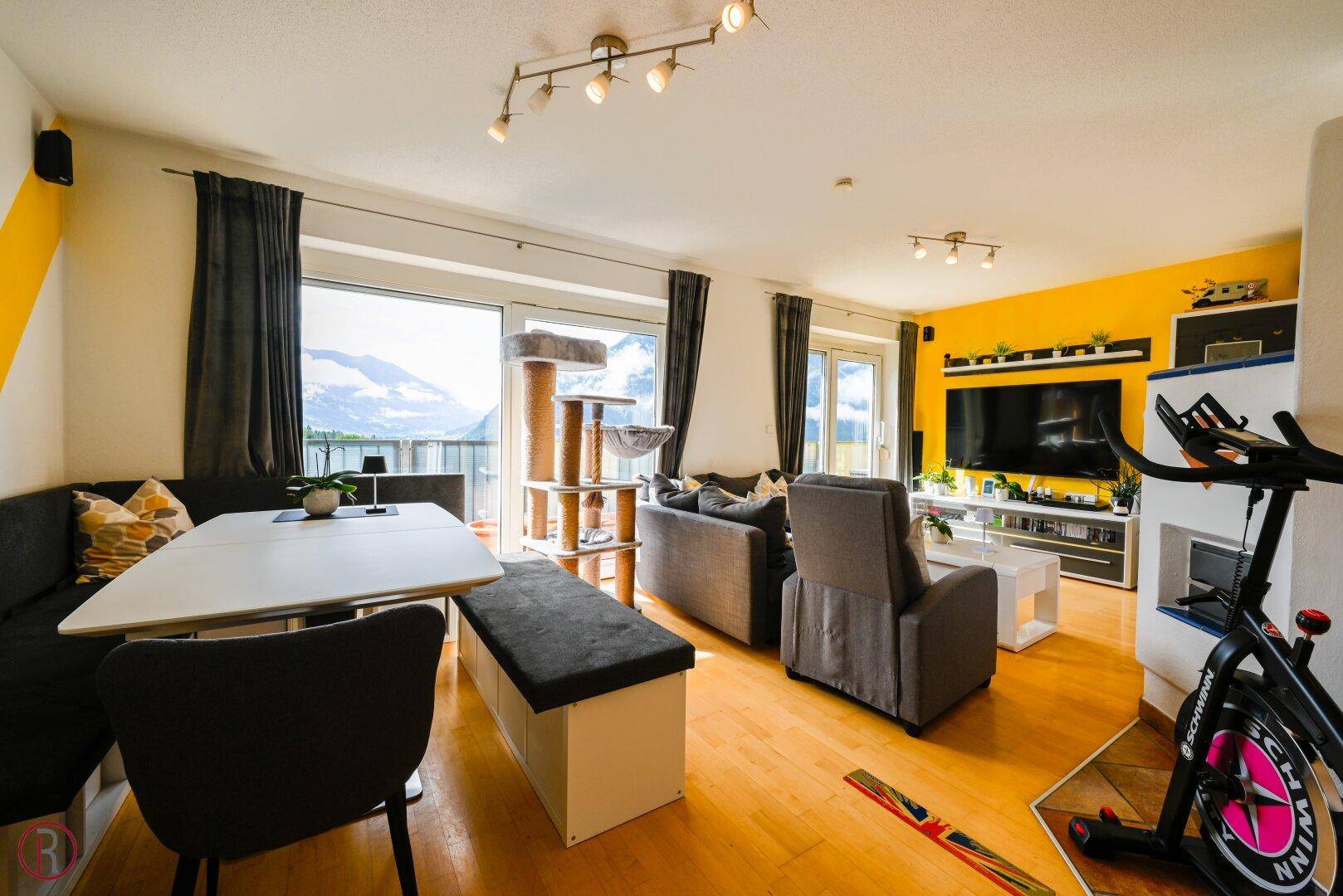 Geräumiger Wohn-Essbereich mit Holzboden, Fitnessgerät und Zugang zum Balkon mit Bergblick.