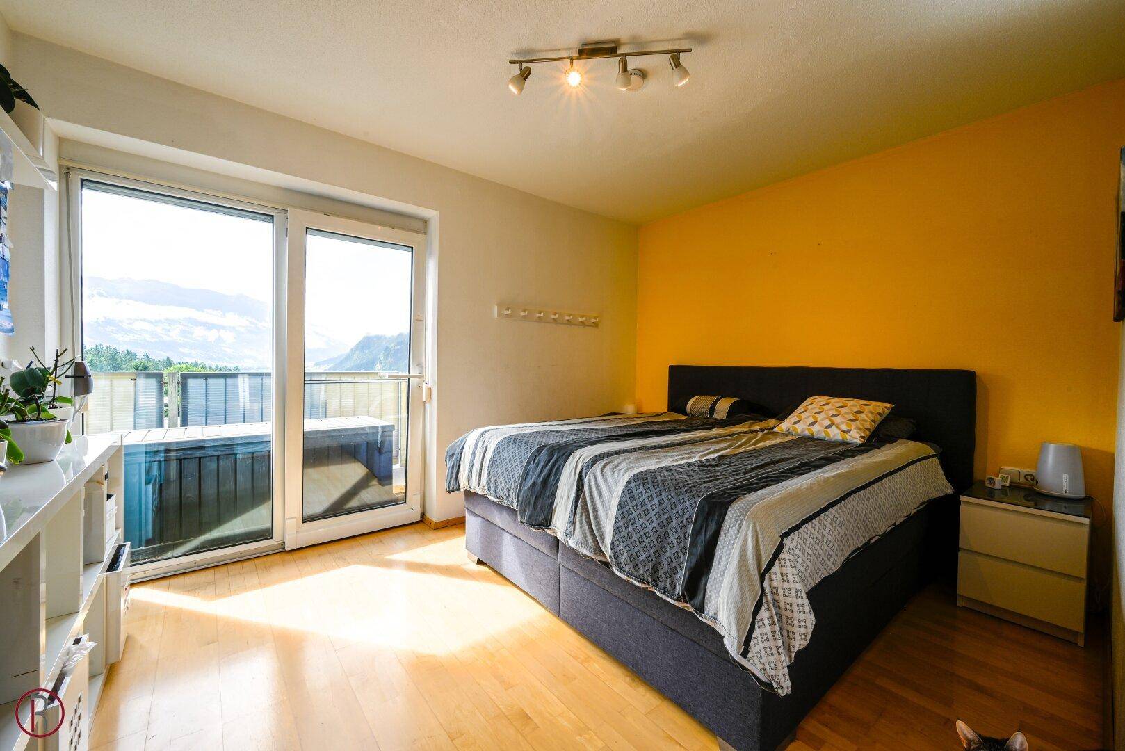 Helles Schlafzimmer mit Doppelbett, gelber Akzentwand und Zugang zu einem Balkon mit Bergblick.