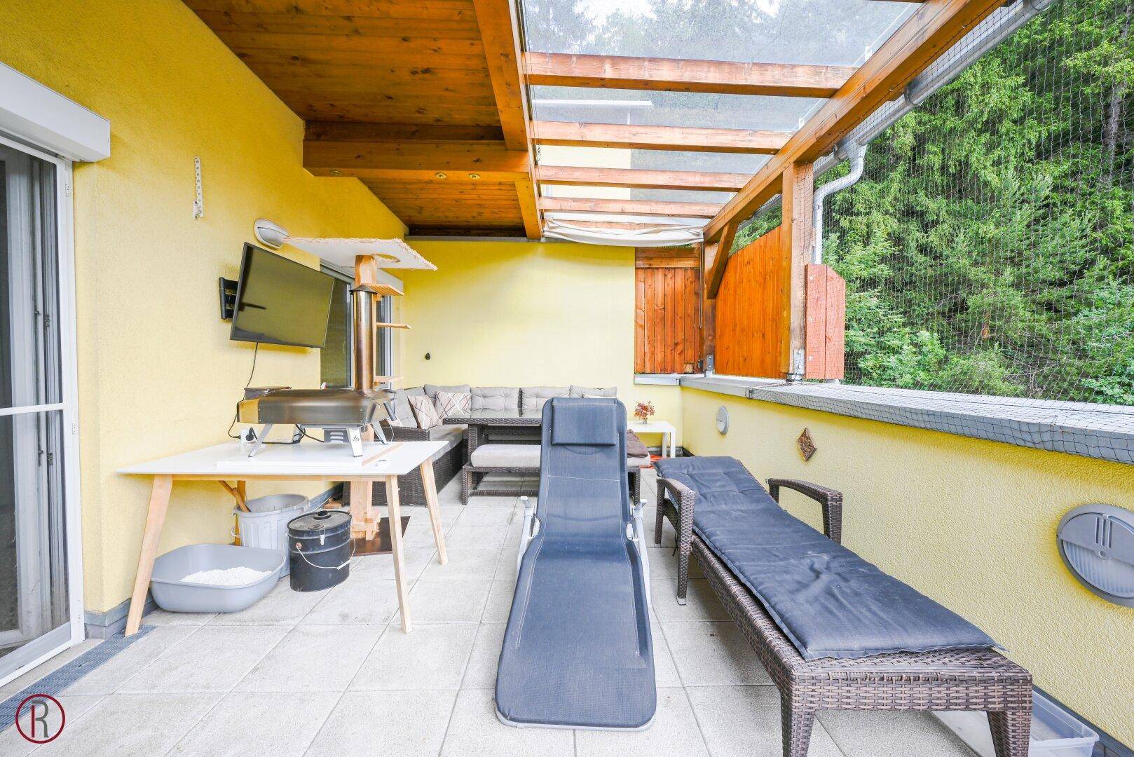 Überdachte Terrasse mit Outdoor-Möbeln, Fernseher und einer privaten Sauna, umgeben von Grün.
