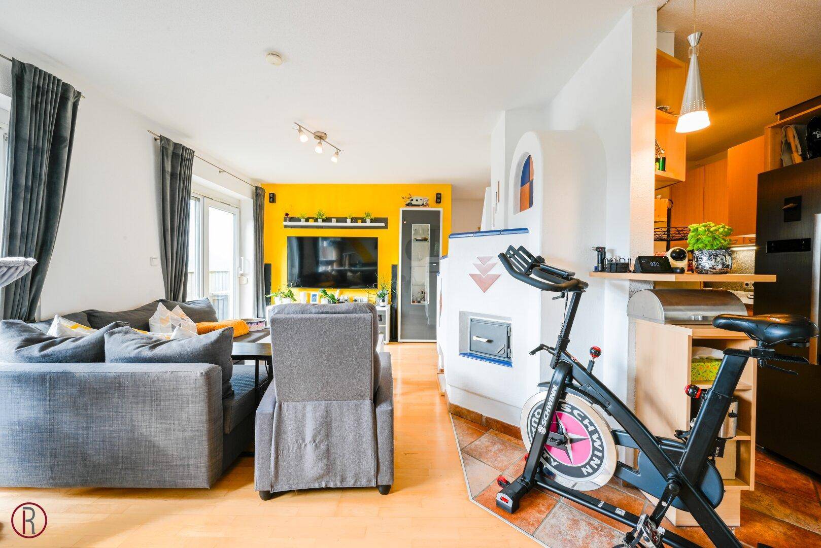 Gemütliches Wohnzimmer mit Kachelofen, Sofa und Fitnessgerät, offener Übergang zur Küche.
