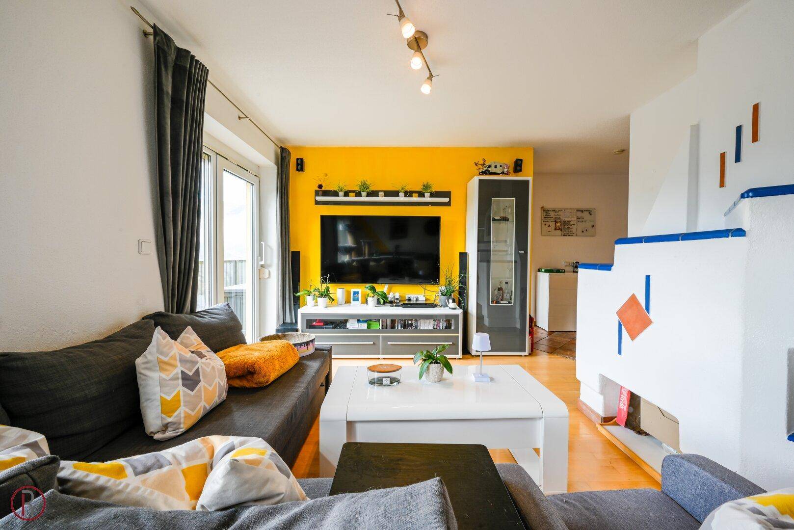 Modernes Wohnzimmer mit gelber Akzentwand, Fernseher und einem dekorativen Kachelofen.