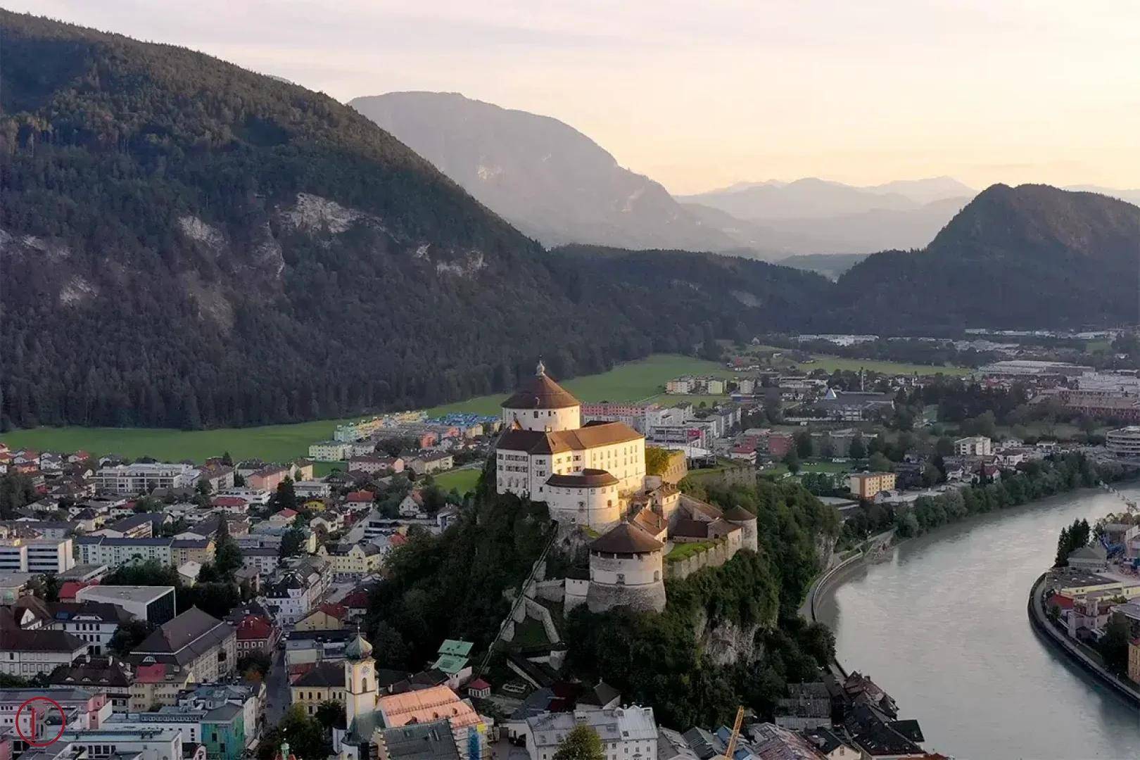 Luftaufnahme einer historischen Burg auf einem Hügel, umgeben von einer Stadt, einem Fluss und majestätischen Bergen.