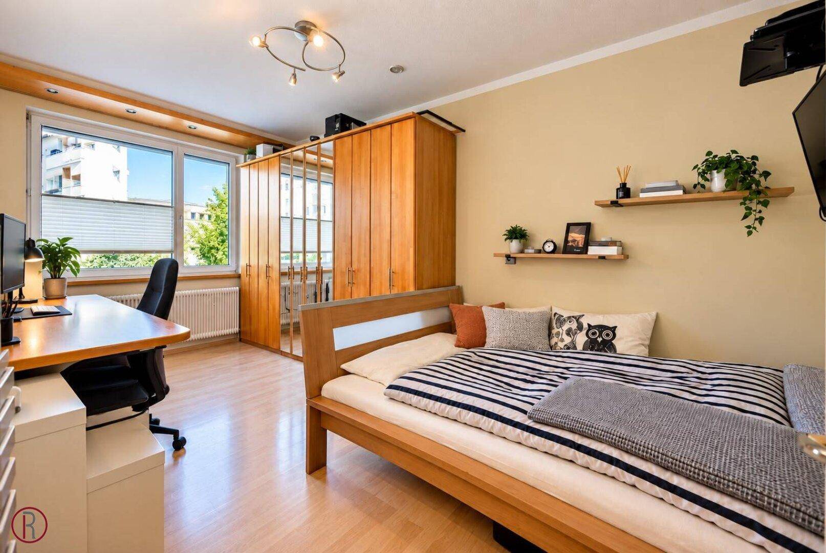 Schlafzimmer mit hellem Holzfußboden, großem Fenster und integriertem Arbeitsbereich.
