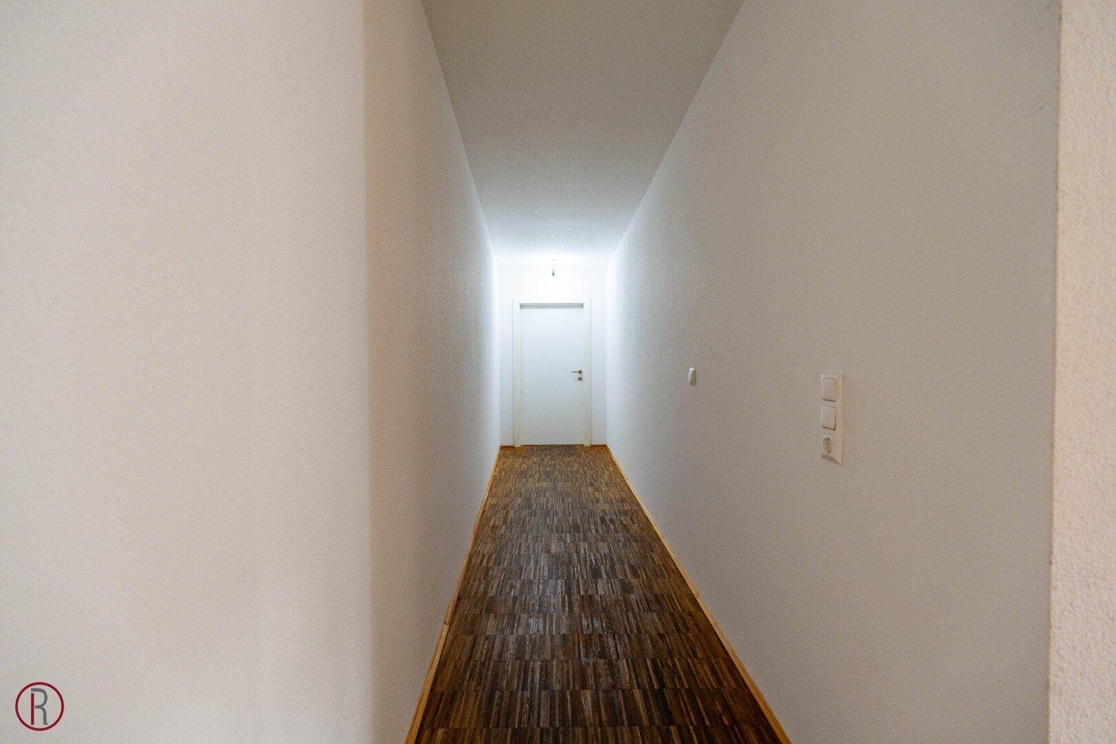 Langer, schmaler Flur mit Holzboden und weißen Wänden, der zu weiteren Räumen führt.