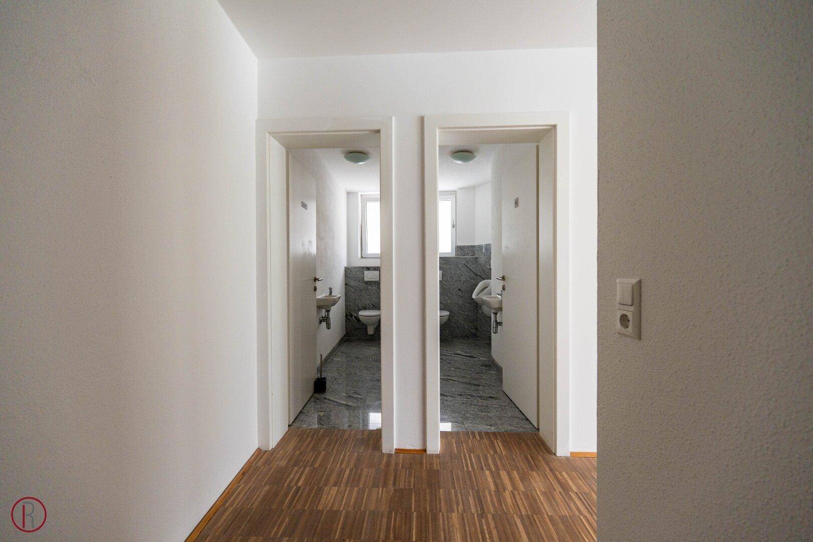 Zwei separate Toilettenräume mit Waschbecken und Spiegeln, zugänglich über einen Flur.