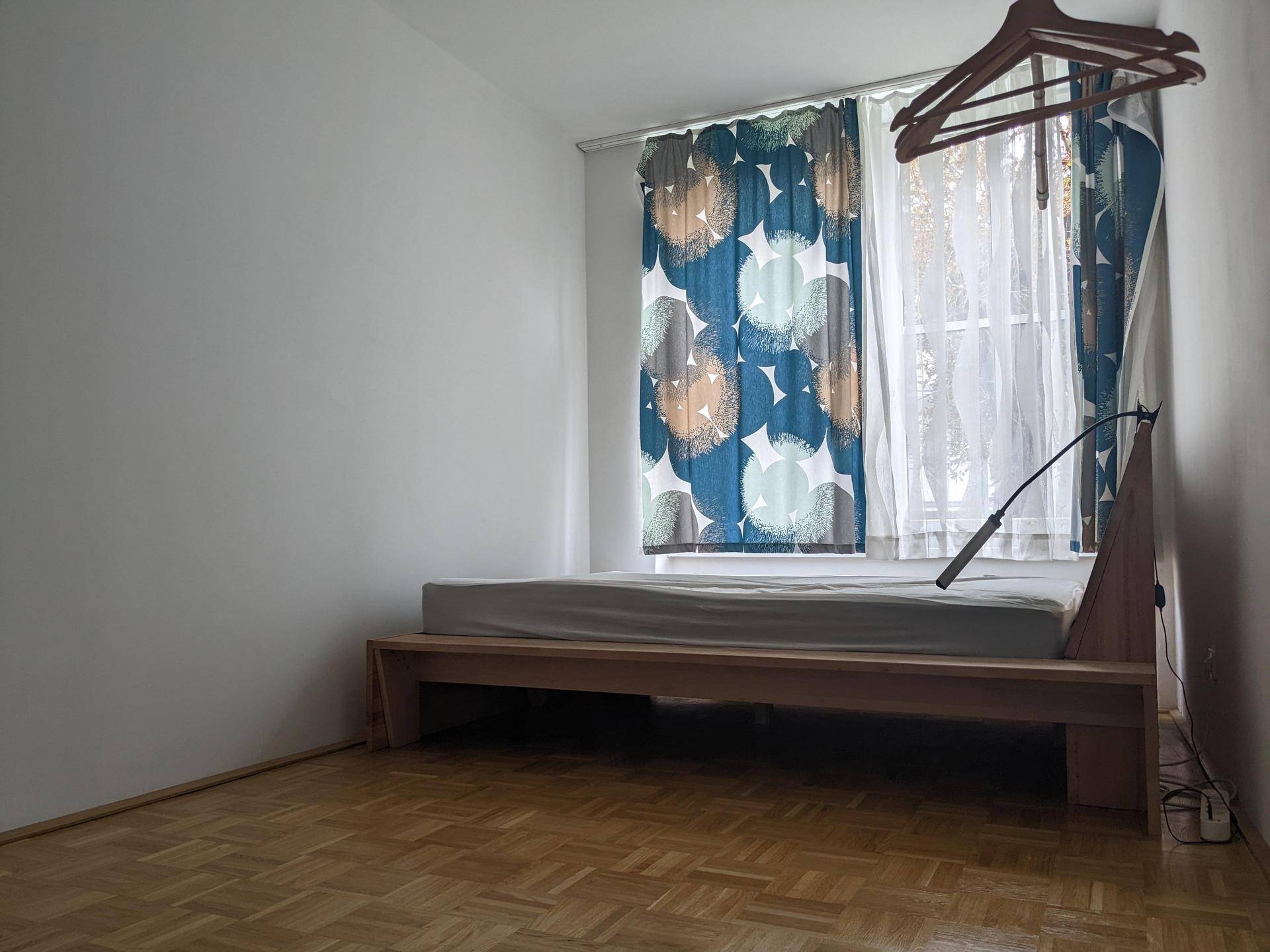Schlafzimmer mit Holzbettgestell, Parkettboden und farbenfrohen Vorhängen vor dem Fenster.