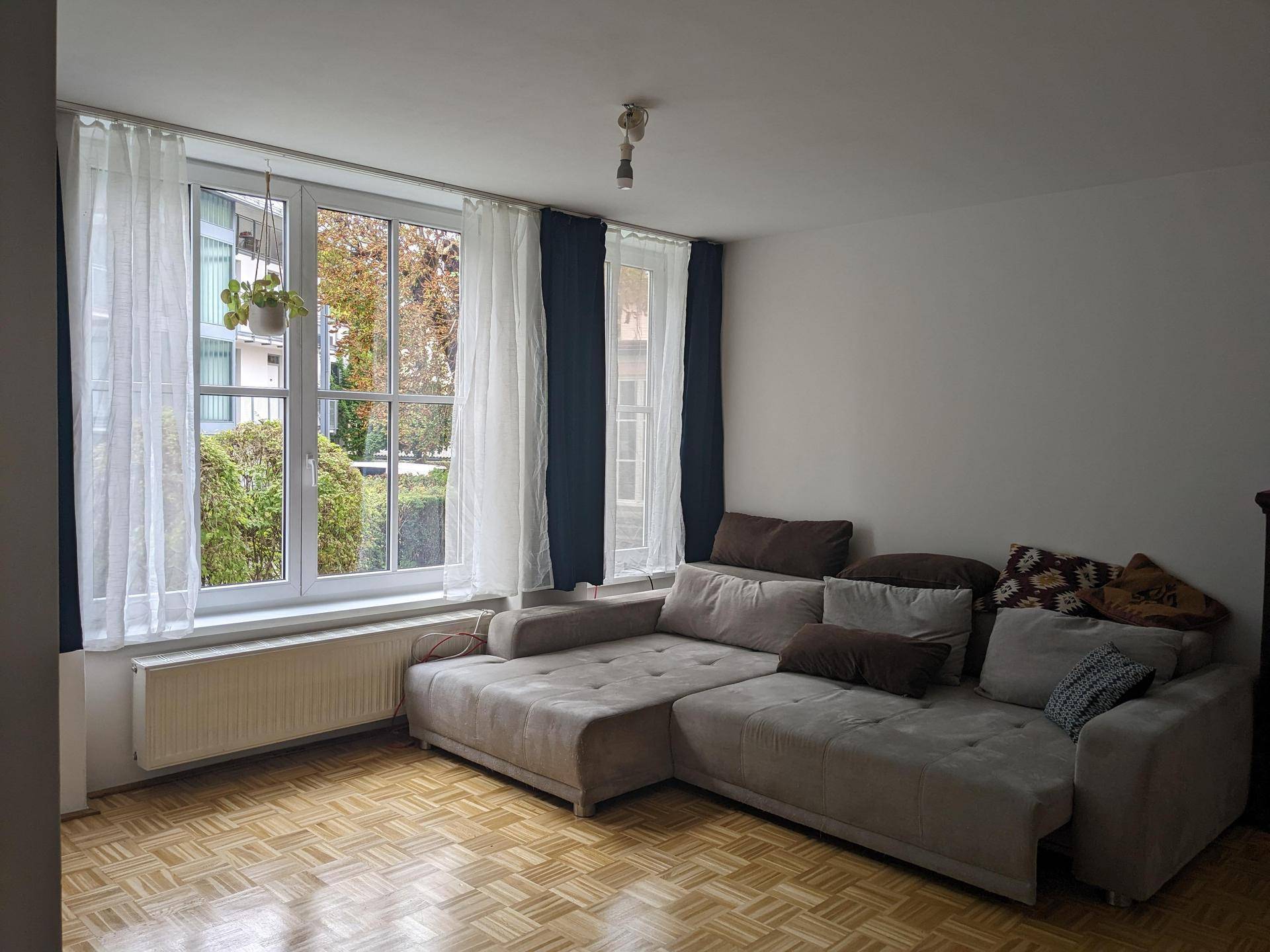 Gemütliches Wohnzimmer mit großem Ecksofa, Parkettboden und hellen Fenstern mit Blick ins Grüne.
