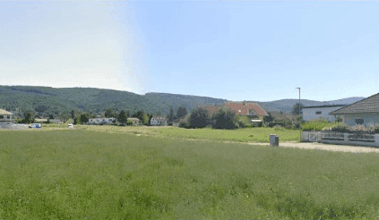 Weitläufige grüne Wiesenfläche mit Blick auf Wohnhäuser und bewaldete Hügel in der Ferne.