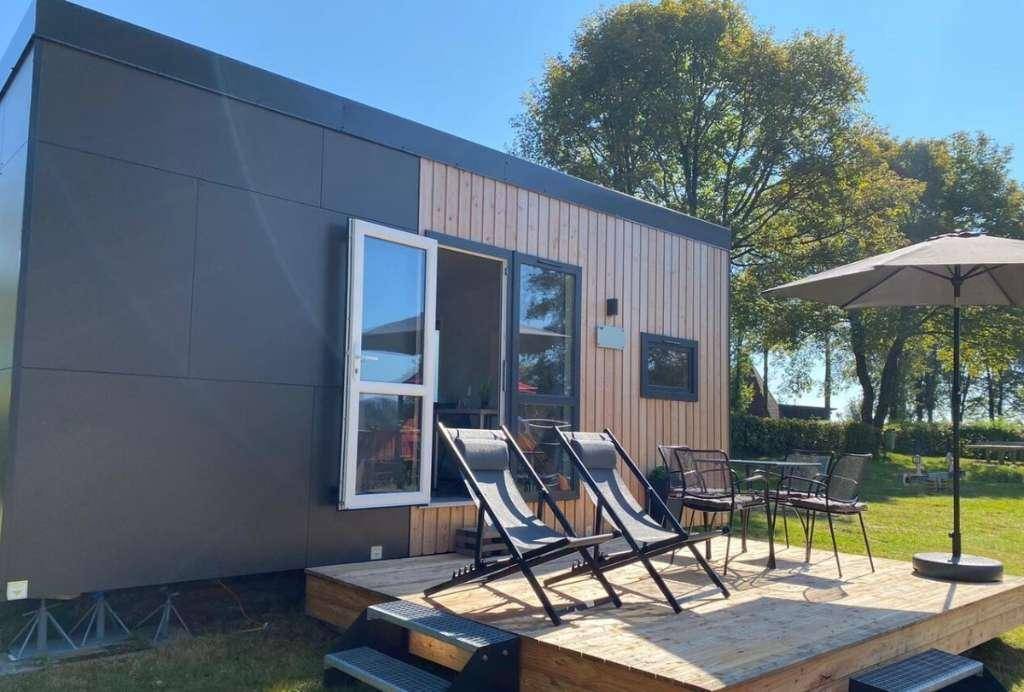 Moderne Tiny-House-Fassade mit Holzverkleidung, kleiner Terrasse und Liegestühlen im Grünen.