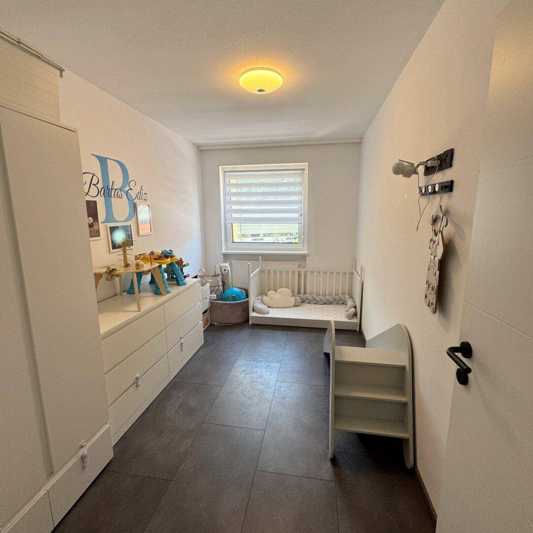Kinderzimmer mit Babybett, weißer Kommode und Spielzeug, ausgestattet mit dunklem Fliesenboden.