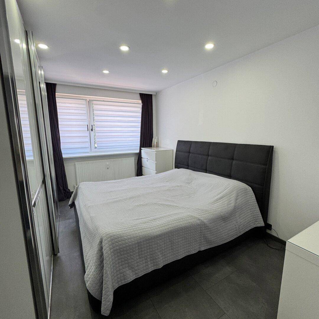 Schlafzimmer mit Doppelbett, großem Spiegelschrank und Fenster mit Jalousien, auf Fliesenboden.