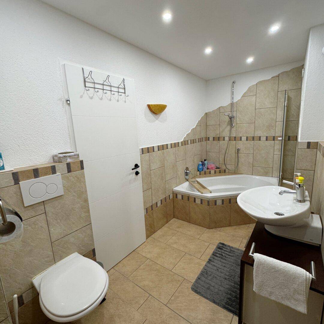 Badezimmer mit beigefarbenen Fliesen, einer Eckbadewanne, Toilette und modernem Waschbecken.
