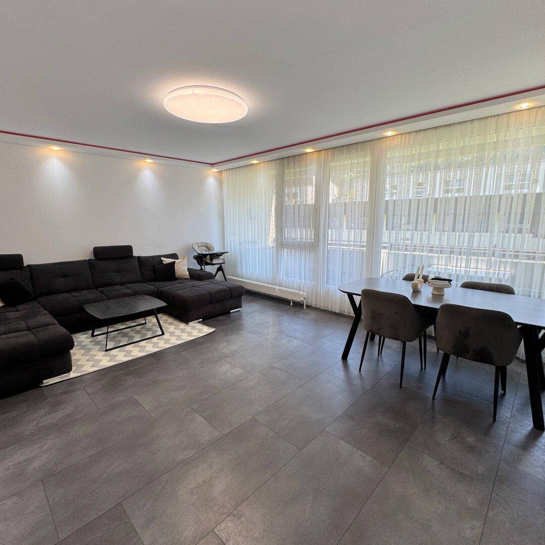 Großzügiger Wohn-Essbereich mit dunklem Sofa, Esstisch und viel natürlichem Licht durch die Fenster.