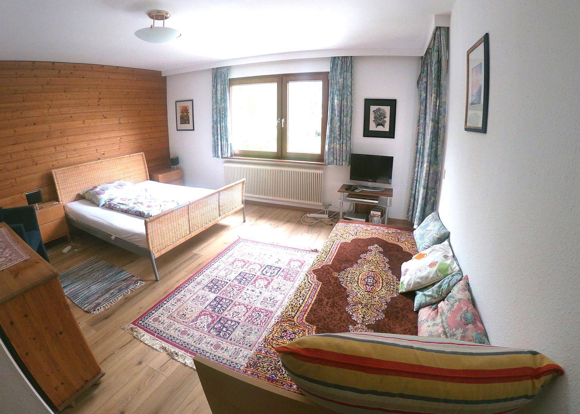 Gemütliches Schlafzimmer mit Holzwandverkleidung, Teppichen und einem bequemen Sofa am Fenster.