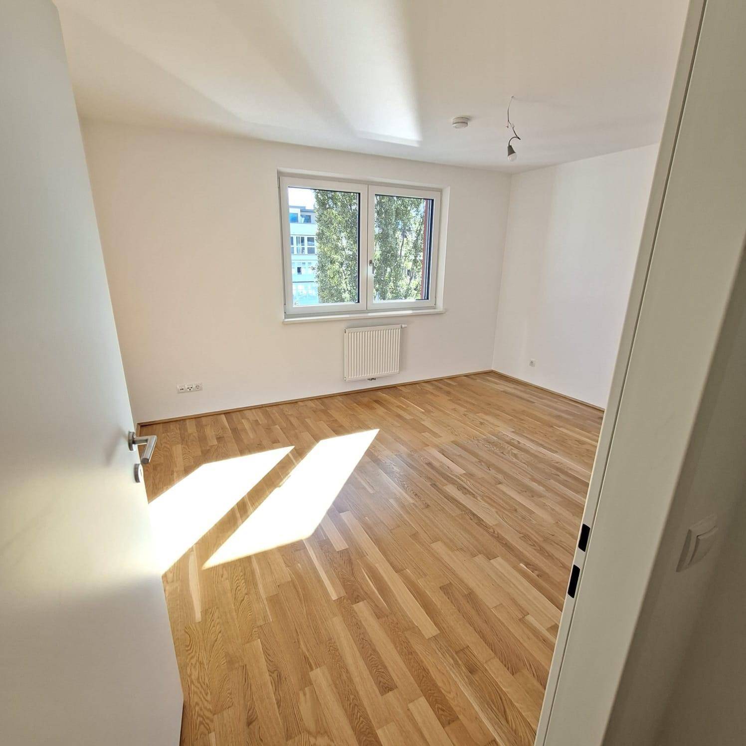 Helles Schlafzimmer mit Parkettboden und großem Fenster, das viel Tageslicht hereinlässt.