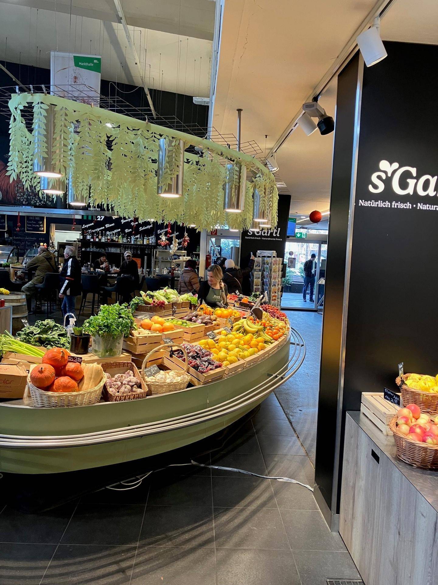 Heller Innenbereich einer Markthalle mit frischem Obst und Gemüse auf einer ansprechenden Boots-Theke präsentiert.