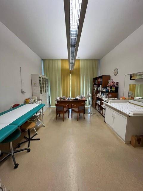 Geräumiges Behandlungszimmer oder Büro mit Untersuchungsliege, Schreibtisch und Bücherregal.