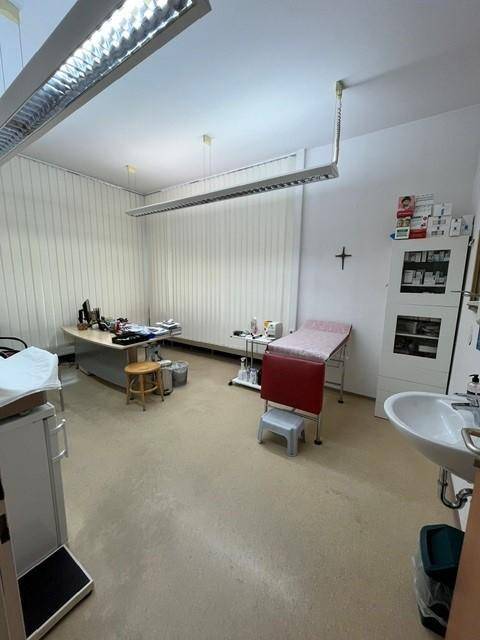 Helles Untersuchungszimmer mit medizinischer Ausstattung, Waschbecken und Jalousien an den Fenstern.