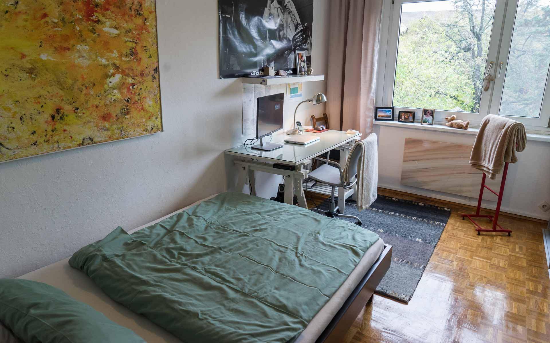 Schlafzimmer mit Bett, Schreibtisch und Fenster, das viel Tageslicht hereinlässt.