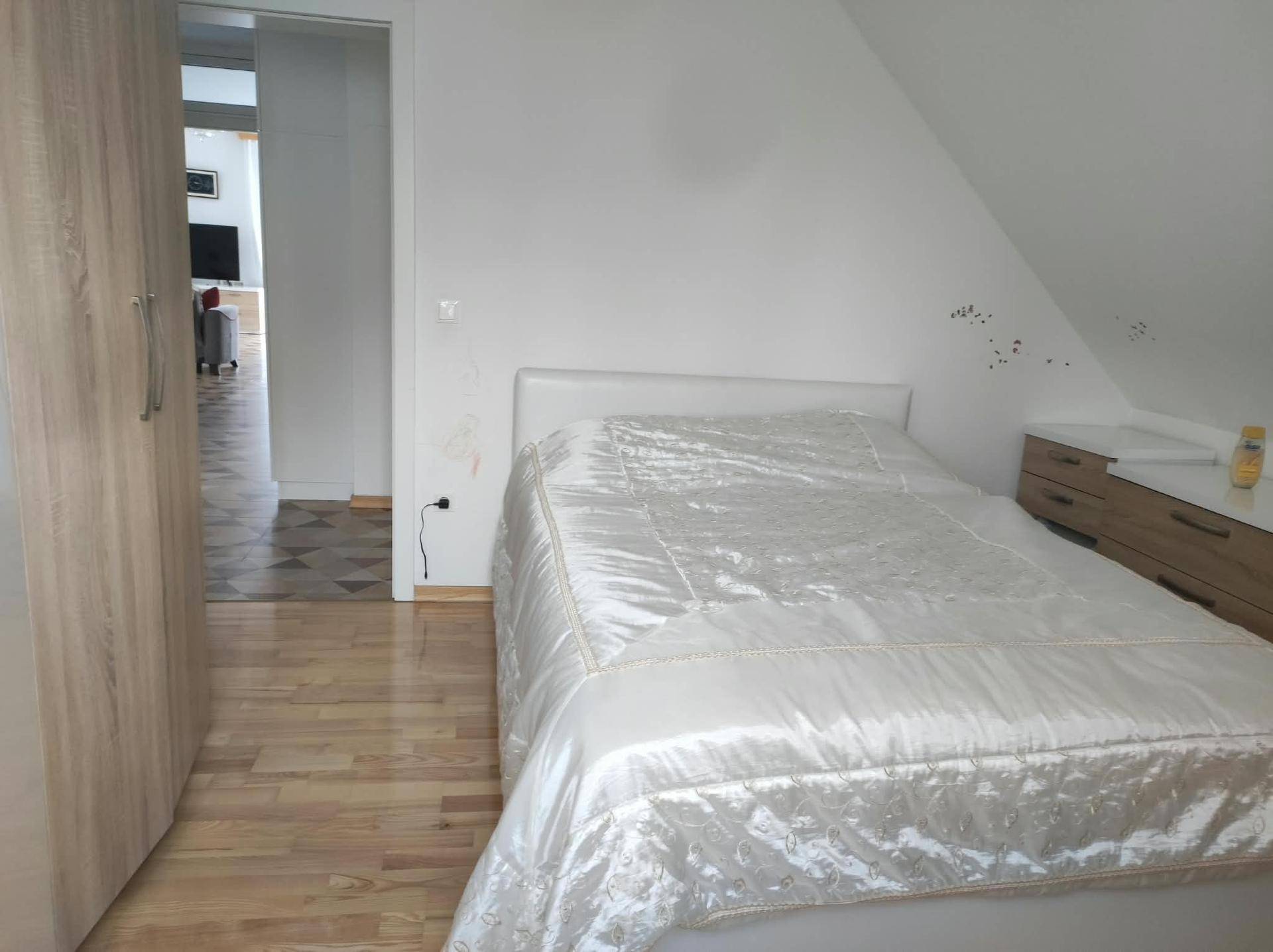 Schlafzimmer mit hellem Holzschrank, weißem Bett und Holzboden, führt zum Flur.