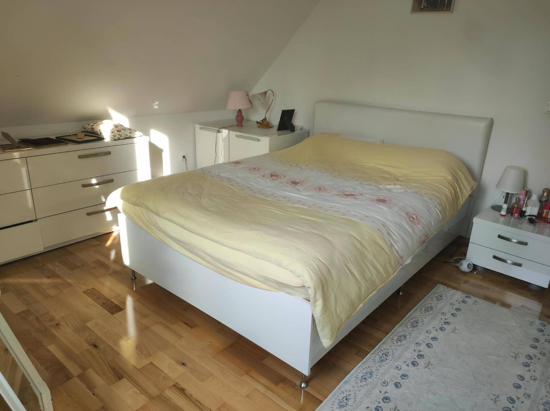 Helles Schlafzimmer mit weißem Bett, Kommode, Holzfußboden und schräger Decke.