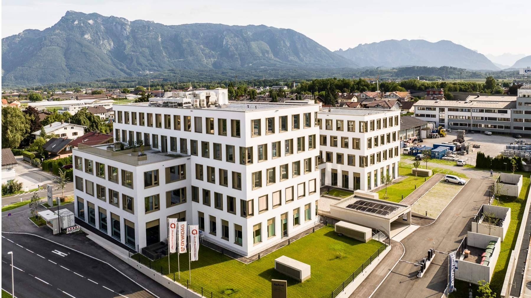 Luftaufnahme eines modernen Gebäudekomplexes mit Parkplätzen und Grünflächen, umgeben von Bergen.