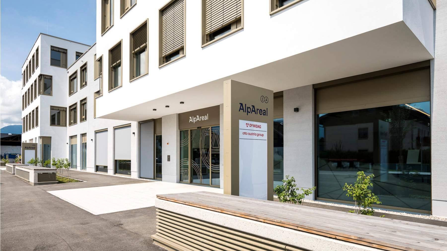 Eingangsbereich des modernen Bürogebäudes mit Glasfassade und Beschilderung 'AlpAreal'.