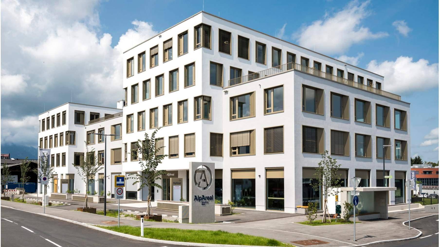 Winkelansicht eines modernen weißen Bürogebäudes mit vielen Fenstern und einem "AlpAreal"-Schild an der Ecke.