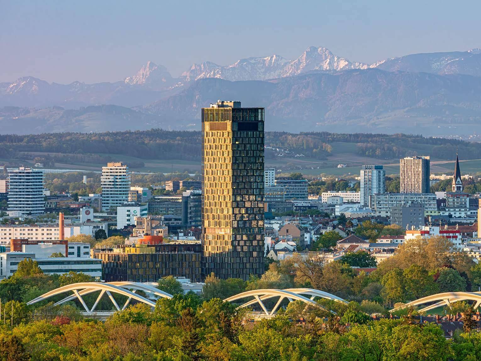 Panoramablick auf eine Stadt mit einem markanten Hochhaus und schneebedeckten Bergen im Hintergrund.