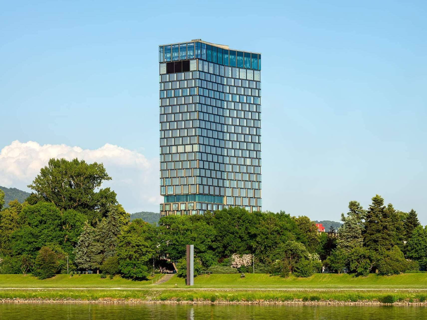 Hohes, modernes Gebäude mit Glasfassade am Ufer eines Gewässers, umgeben von Bäumen.