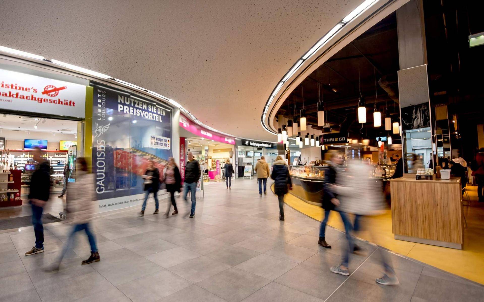 Belebter Gang in einem Einkaufszentrum mit verschiedenen Geschäften und moderner Beleuchtung.