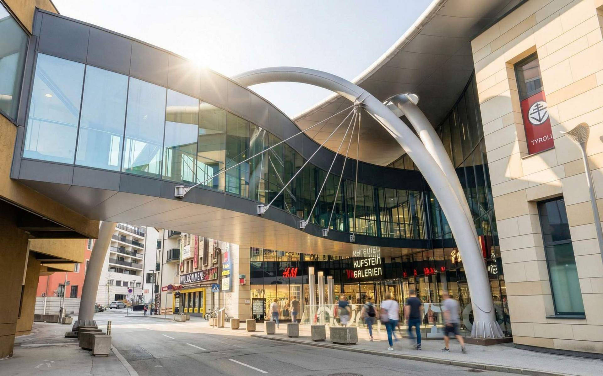 Moderne Architektur eines Einkaufszentrums mit Glasfassade und Fußgängerbrücke bei Sonnenschein.