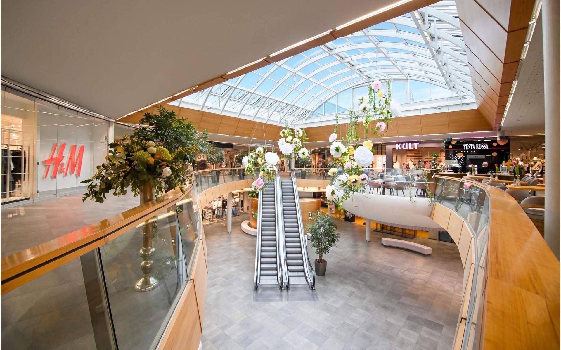 Helles Einkaufszentrum-Atrium mit Rolltreppen, Glasdach und dekorativen Hängeelementen.