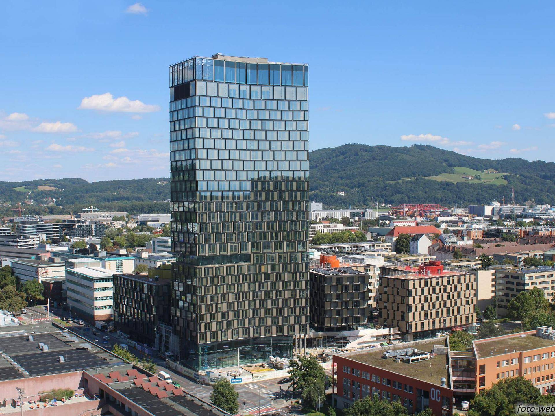 Panoramablick auf den modernen Gebäudekomplex und die umliegende Stadtlandschaft mit Hügeln im Hintergrund.