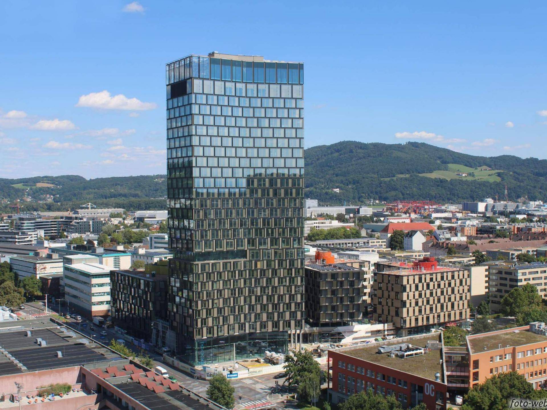 Ansicht eines modernen Hochhauses mit Glasfassade, umgeben von städtischer Bebauung und grünen Hügeln im Hintergrund.